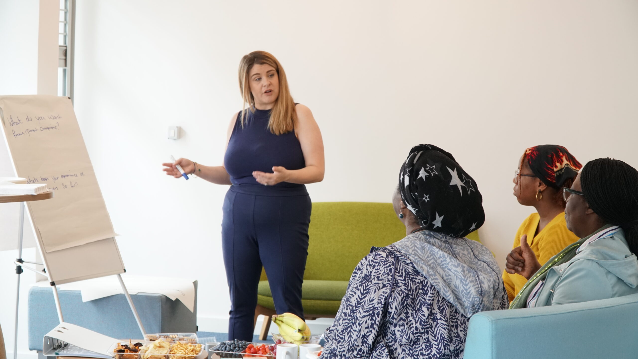 a blonde lady speaks to a crowd, gesturing toward a whiteboard